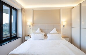 Quarto de hotel com uma cama grande, duas mesas de cabeceira, uma janela com cortinas e luminárias fixadas na parede.