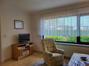 Sala de estar com televisão, armário de madeira, poltrona e janela com cortinas de renda.