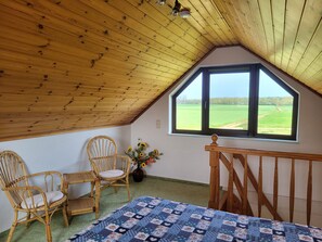 A room with a sloped ceiling, a large window, two wicker chairs, a wooden table, and a quilted bedspread.
