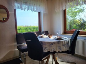 A dining table with chairs, a view of greenery outside, and a window with lace curtains.
