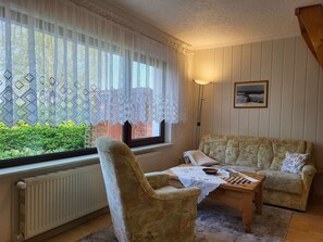 Une pièce chaleureuse avec un fauteuil à motifs, un canapé, une table d’échecs et une vue à travers des rideaux en dentelle.