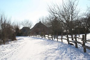 A snow-covered path leading to a building with a thatched roof, surrounded by bare trees and a wooden fence.