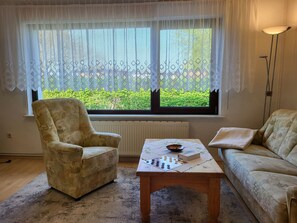Una habitación con un ventanal grande, un radiador, una mesa de madera con un juego de mesa, un sillón estampado y un sofá.