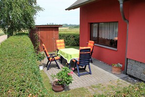 Um edifício vermelho com fachada de tijolos, porta de madeira e janela com cortinas de renda. Há uma mesa com toalha verde e duas cadeiras com almofadas laranja e azuis. Um vaso com flores cor-de-rosa está em frente à mesa.