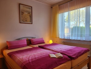 Uma cama de casal com roupa de cama rosa, um abajur, um quadro na parede e uma janela com cortinas leves.