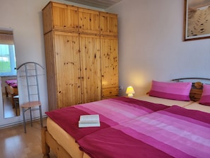 Un dormitorio con un armario de madera, una cama con ropa de cama rosa, una lámpara de noche y una ventana con vistas al exterior.