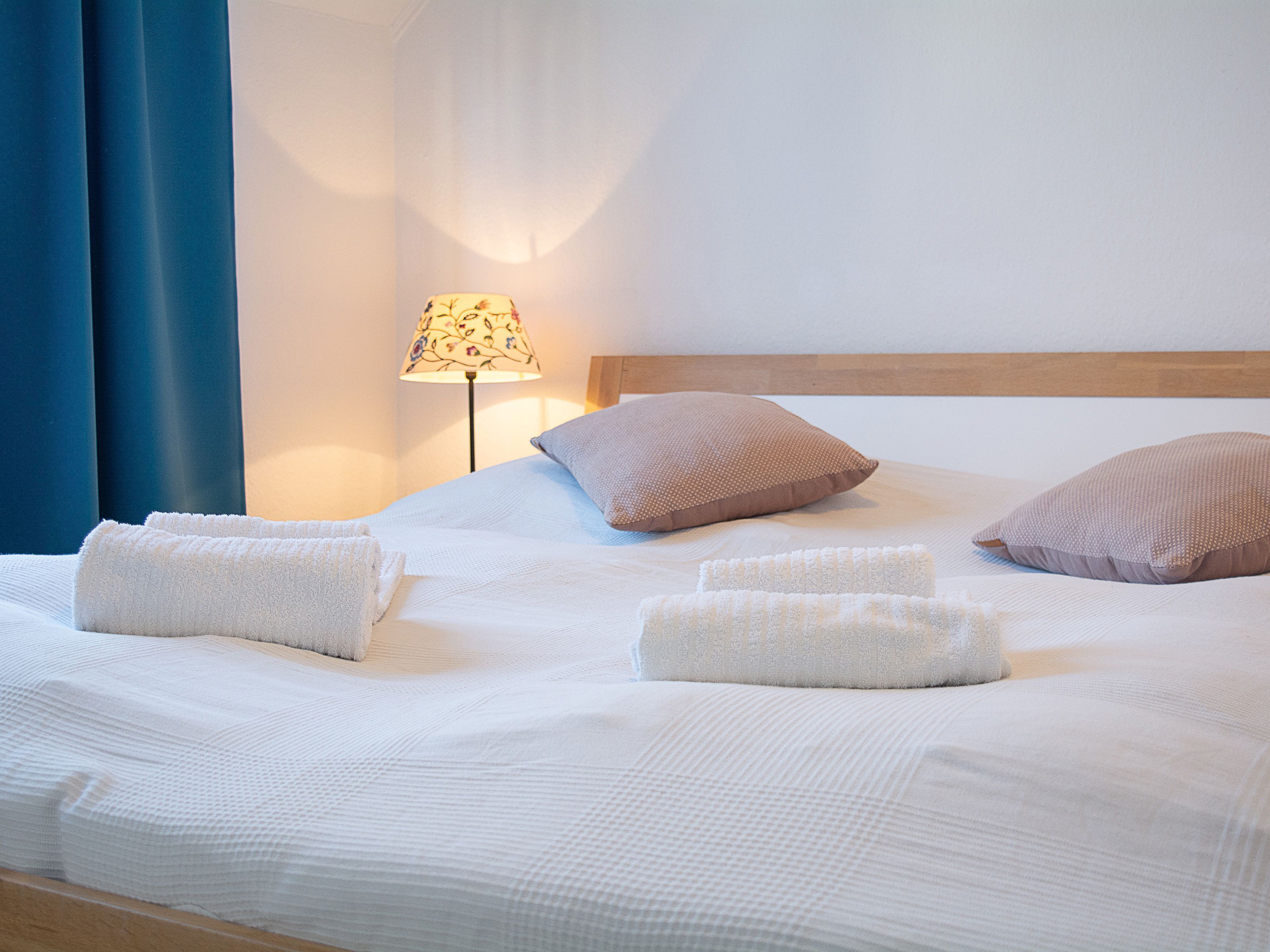 A neatly made bed with white linens and two pillows, a lamp with a floral shade, and a blue curtain.