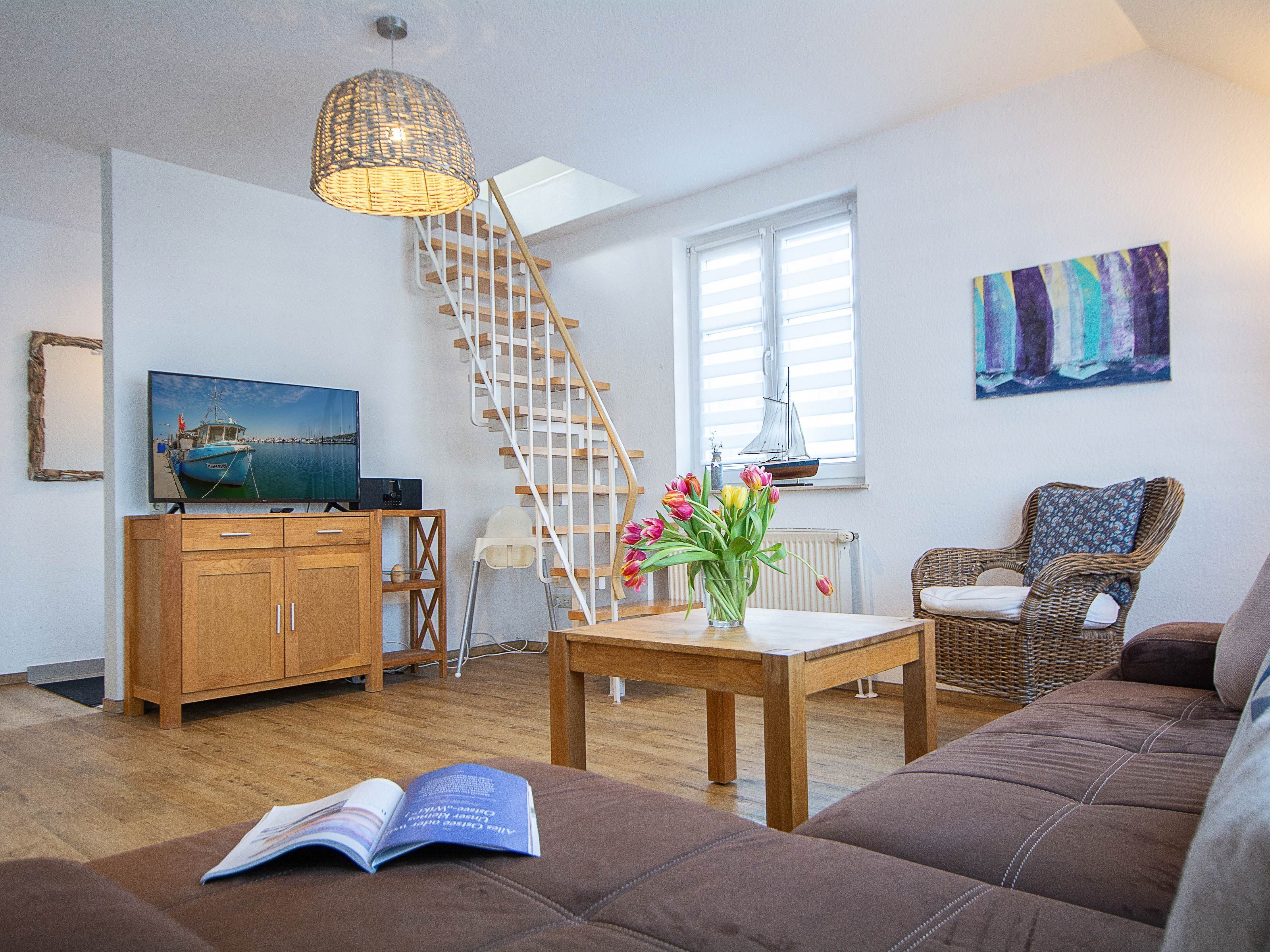 Een woonkamer met een houten trap, een televisie, een boekenkast, een bijzettafeltje met bloemen, een rieten stoel en een bruine bank.