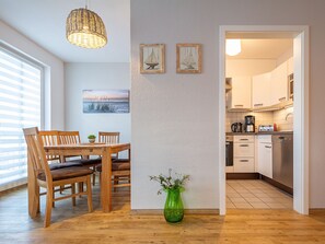 Un coin repas avec une table et des chaises en bois, un vase vert rempli de fleurs et un tableau représentant un voilier accroché au mur.
