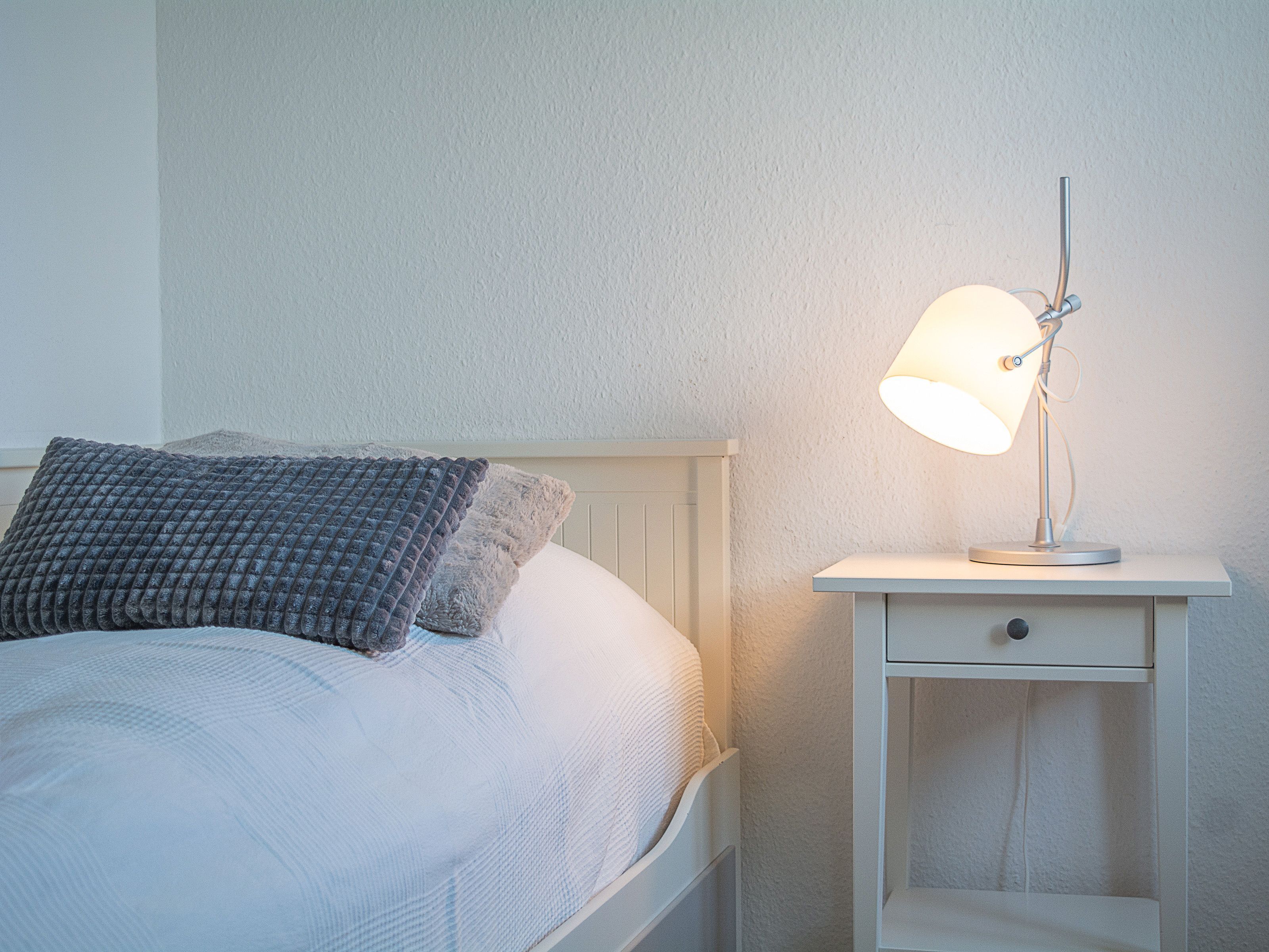 A neatly made bed with a white headboard and a gray textured pillow. A white nightstand with a lamp and a drawer is beside the bed.