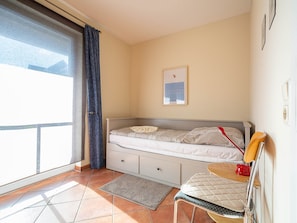 Un dormitorio con cama, una silla, una mesita, una ventana con cortinas y un cuadro colgado en la pared.