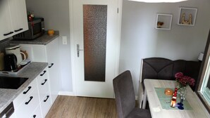 A compact kitchen with a microwave, coffee maker, and orange on the counter. There is a dining area with a table, chairs, and a vase with flowers. - FeWo Weber - GM 69911 - FeWo Weber (Graal-Müritz)
