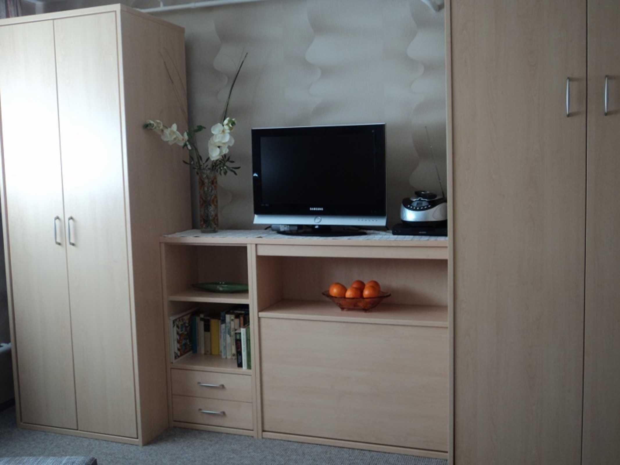 Ein Zimmer mit einem Holztisch, einem Fernseher, einer Vase mit Blumen und einer Orangenschale.
