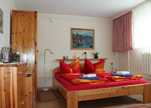 Una cama de madera con ropa de cama roja y naranja, un armario de madera, una lámpara de pie, un cuadro de un paisaje urbano y un radiador.