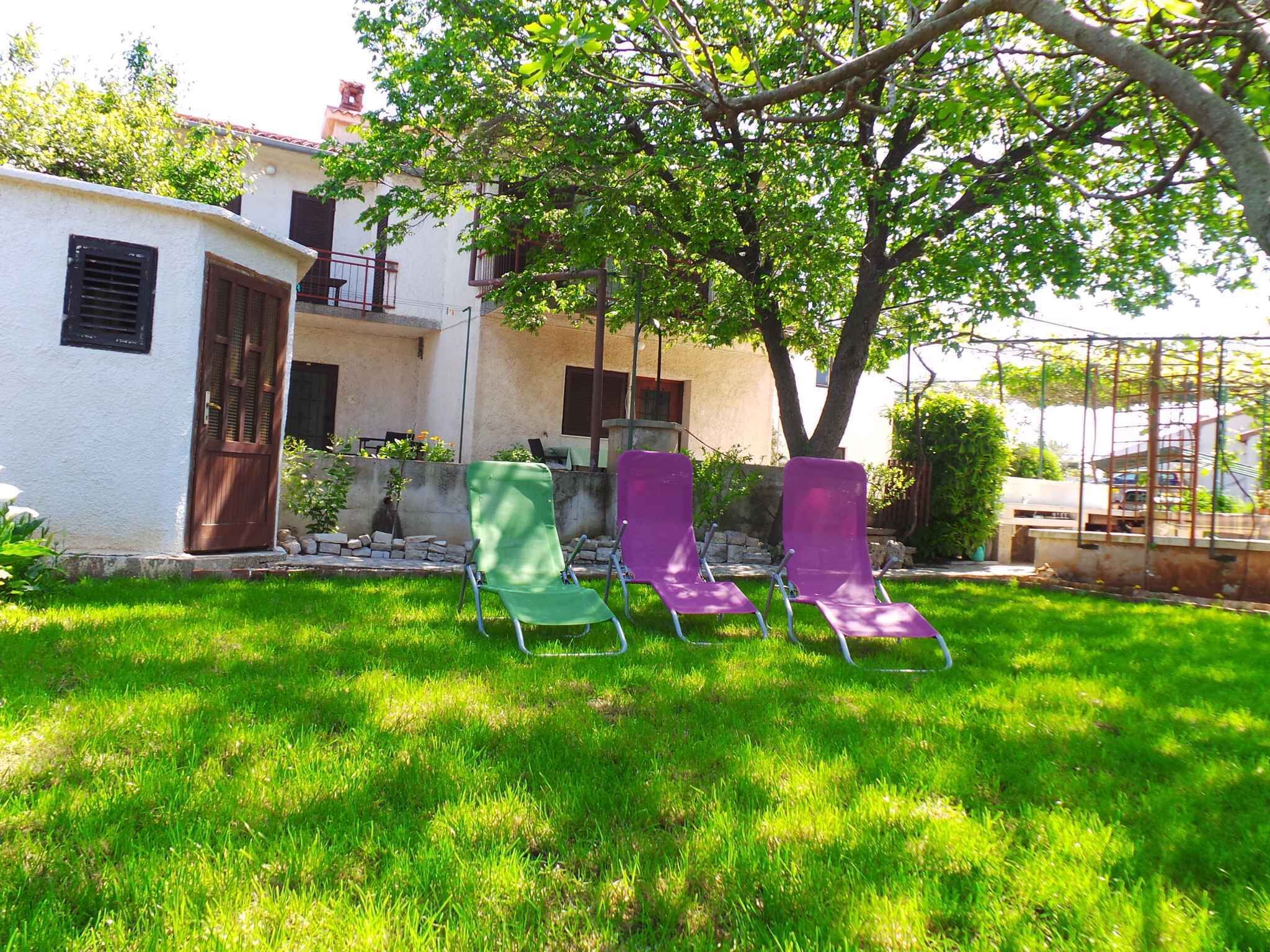 Ein Garten mit einem weißen Haus, grünen und violetten Stühlen und einem Baum, der Schatten spendet.