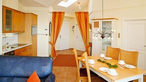 A modern kitchen with wooden cabinets, a dining area with a table set for two, and a blue sofa with an orange pillow. - (60) 3-room apartment with balcony - direct lake view - apartment house (Kühlungsborn)