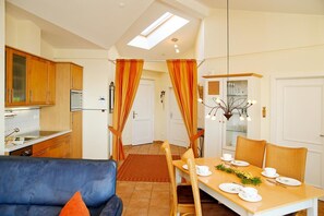 A modern kitchen with wooden cabinets, a dining area with a table set for two, and a blue sofa with an orange pillow. - (60) 3-room apartment with balcony - direct lake view - apartment house (Kühlungsborn)