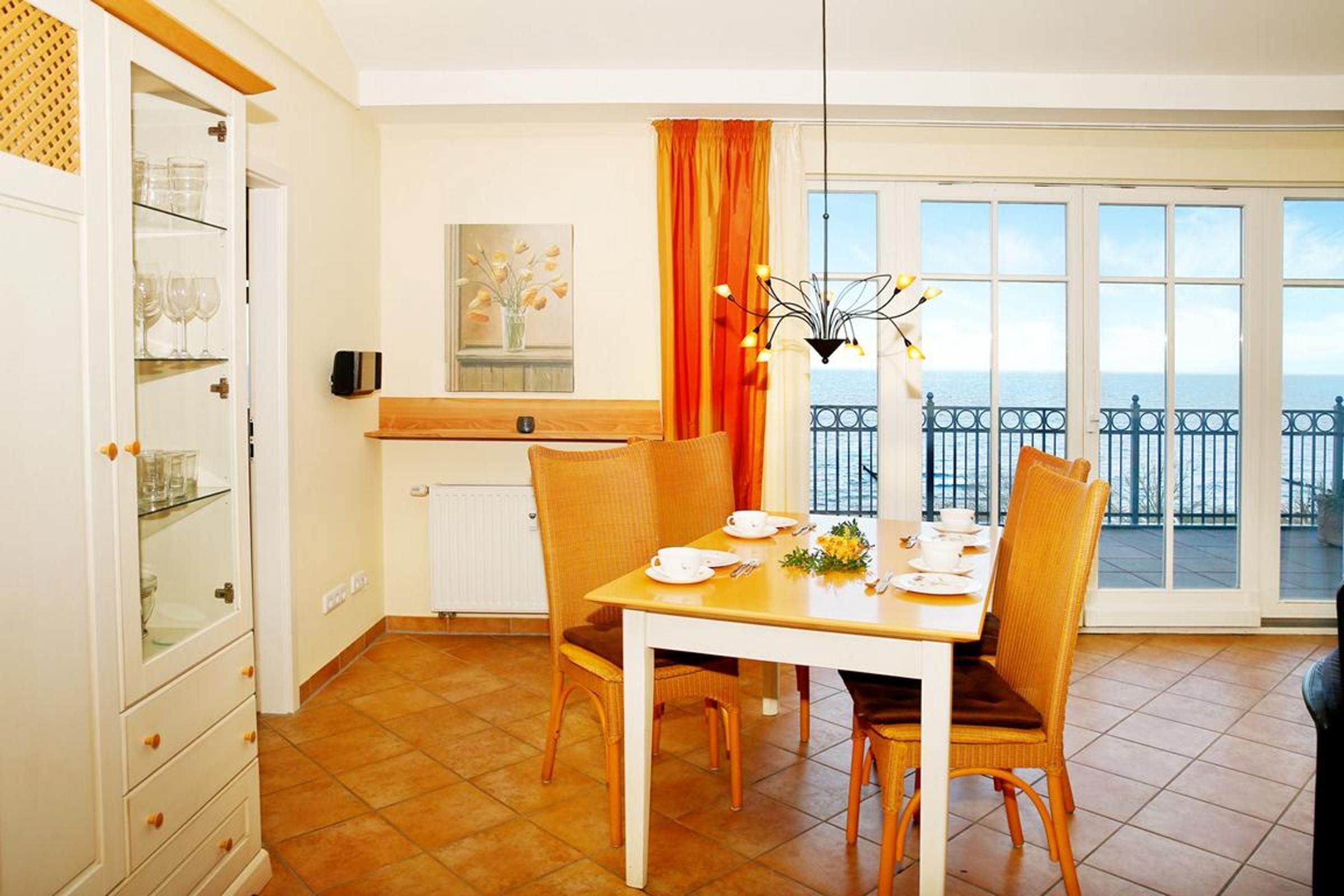 A dining area with a table set for four, a view of the sea through large windows, and a kitchen area with a white cabinet and a radiator.