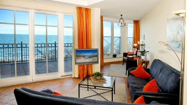 A spacious living room with a large window overlooking the sea, a black sofa with orange cushions, and a television on a glass coffee table. - (60) 3-room apartment with balcony - direct lake view - apartment house (Kühlungsborn)