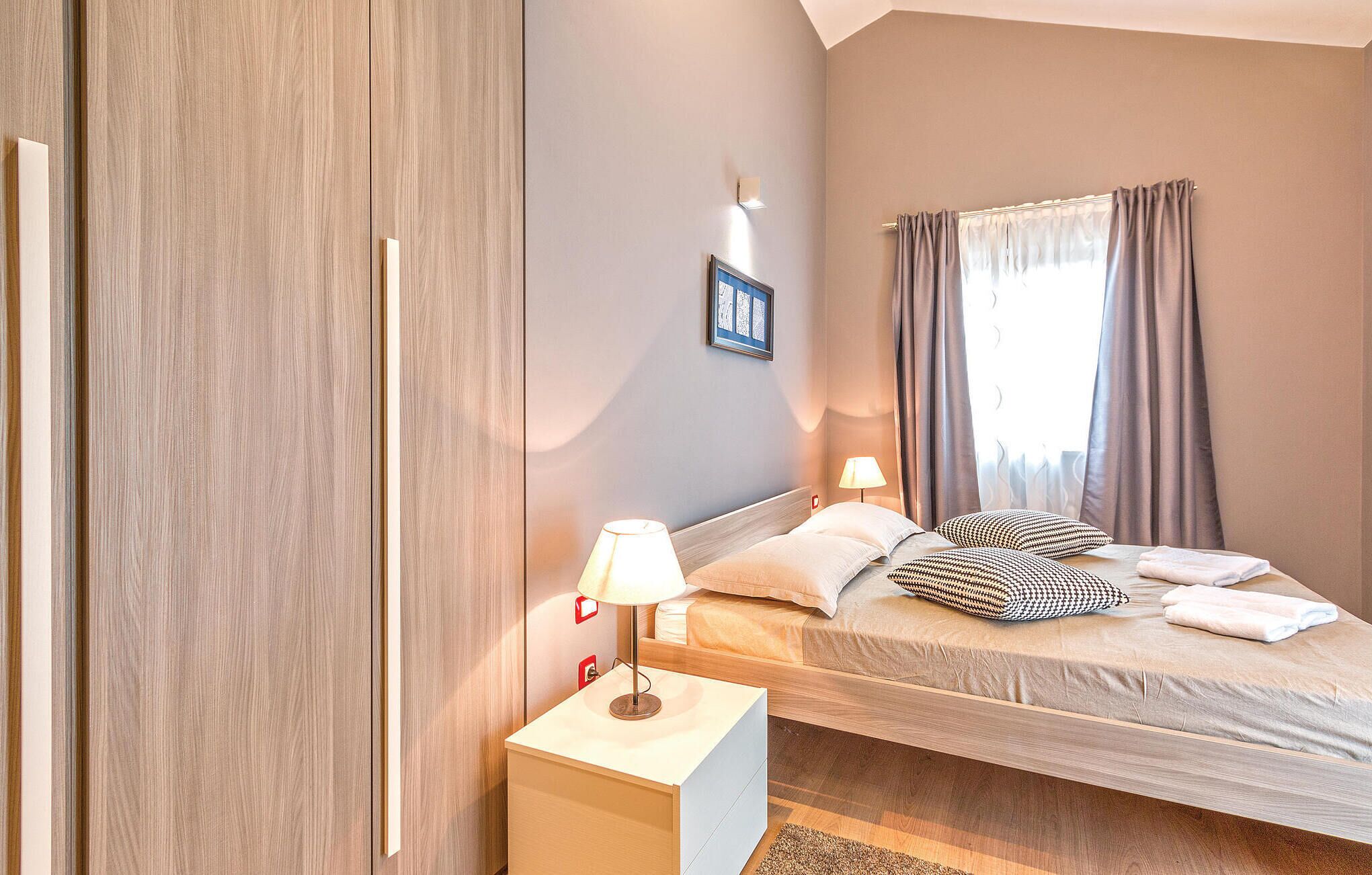 A bedroom with a bed, bedside tables, a nightstand, a wall-mounted lamp, and a window with curtains.