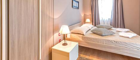A bedroom with a bed, bedside tables, a nightstand, a wall-mounted lamp, and a window with curtains.