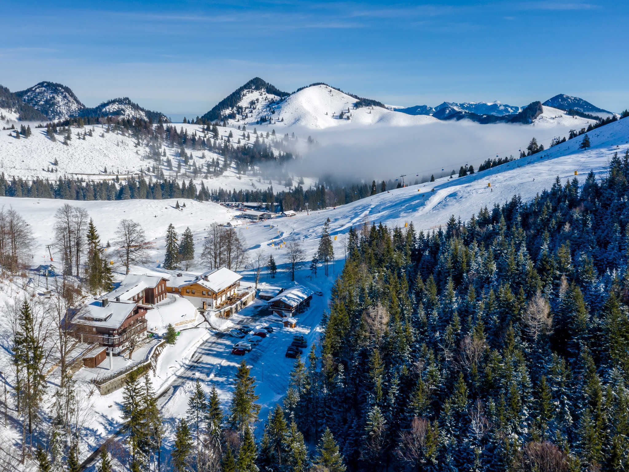 Una località montana innevata con una seggiovia, un'area di parcheggio e un edificio con tetto imbiancato dalla neve.