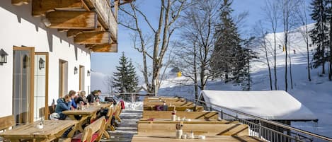 An outdoor wooden deck with benches and tables, a building with a sloped roof, and a snowy landscape with trees and mountains in the background.