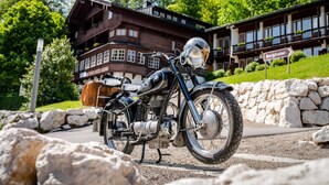 Een klassieke motorfiets geparkeerd voor een groot, meer verdiepingen tellend gebouw met een bord waarop staat 'Berghotel'.
