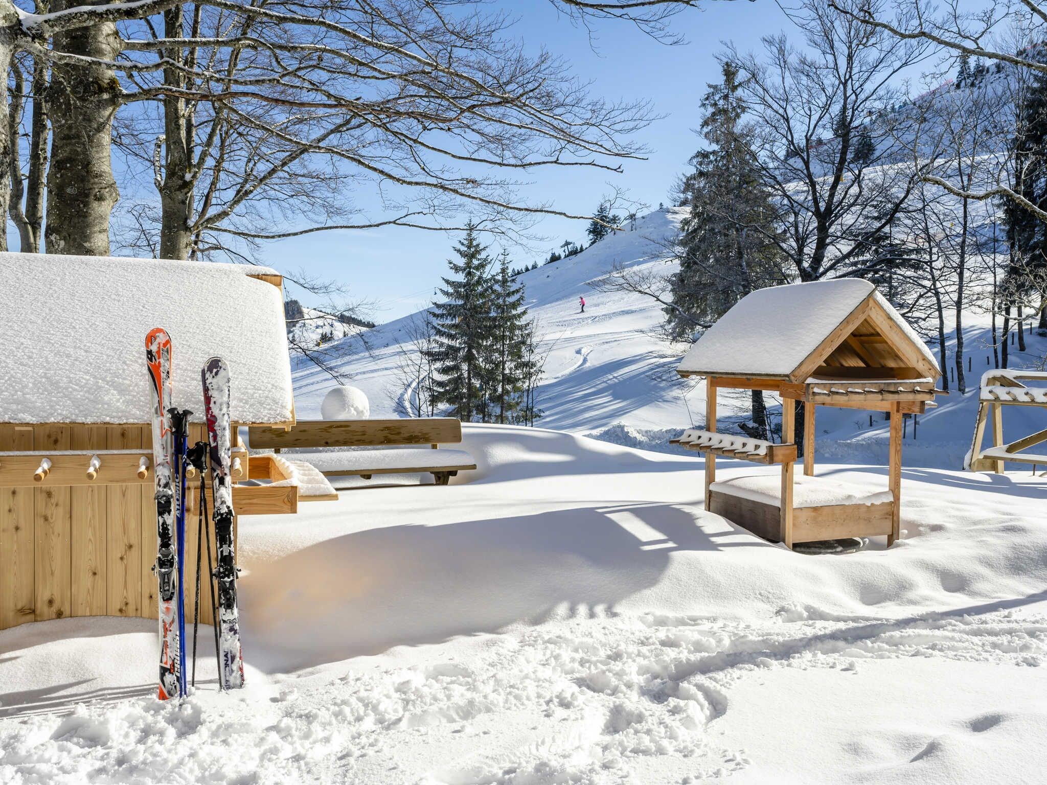 A snow-covered outdoor area with wooden benches and a small wooden structure, likely a shelter or kiosk, with skis leaning against a wooden post.
