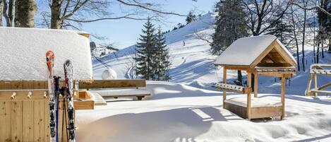 A snow-covered outdoor area with wooden benches and a small wooden structure, likely a shelter or kiosk, with skis leaning against a wooden post.