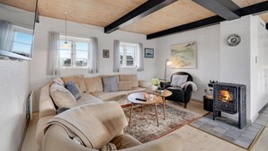 Un salon chaleureux avec un grand canapé d’angle beige, un fauteuil en cuir noir, une petite table ronde ornée d’un vase et d’une bougie, et un poêle à bois.