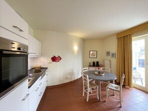 A compact kitchen with a built-in oven, white cabinets, a dining table with chairs, and a large window with curtains.