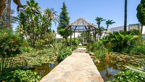 A stone pathway leading to a thatched gazebo surrounded by lush tropical vegetation and a pond with lily pads.