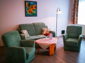 Een woonkamer met een groene zithoek, een bijzettafeltje met een bloevas, een vloerlamp en een schilderij aan de muur.