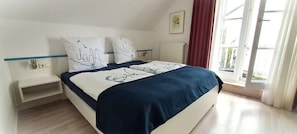 A bedroom with a bed, two pillows, a nightstand, a window with curtains, and a radiator.