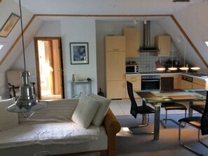 Une chambre à coucher confortable, aménagée dans un style « loft », avec un lit, un bureau et une chaise. On aperçoit, au fond, un coin cuisine équipé de placards et d’un four à micro-ondes.