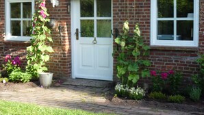 A brick house with a white door and two windows. There are potted plants and flowers in front of the house. - Marc Chagall apartment - Rosengut Falkenstein (Braderup)