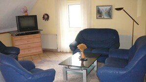 A living room with a television on a wooden cabinet, a coffee table with a vase, two blue armchairs, a floor lamp, and a painting on the wall.
