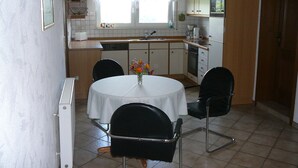 A compact kitchen with a dining area, featuring a round table with chairs, a hanging light fixture, and a window with curtains.