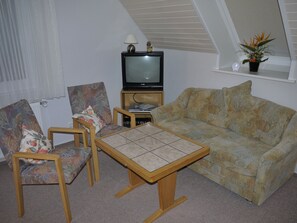 Una sala de estar con un sofá, dos sillones, una mesita, un televisor y una planta en un estante.