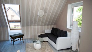 A modern loft bedroom with a sloped ceiling, a white sectional sofa, a glass coffee table, a bench, and a floor lamp. - Apartment Lischewski - Volker Lischewski (Steinberg)
