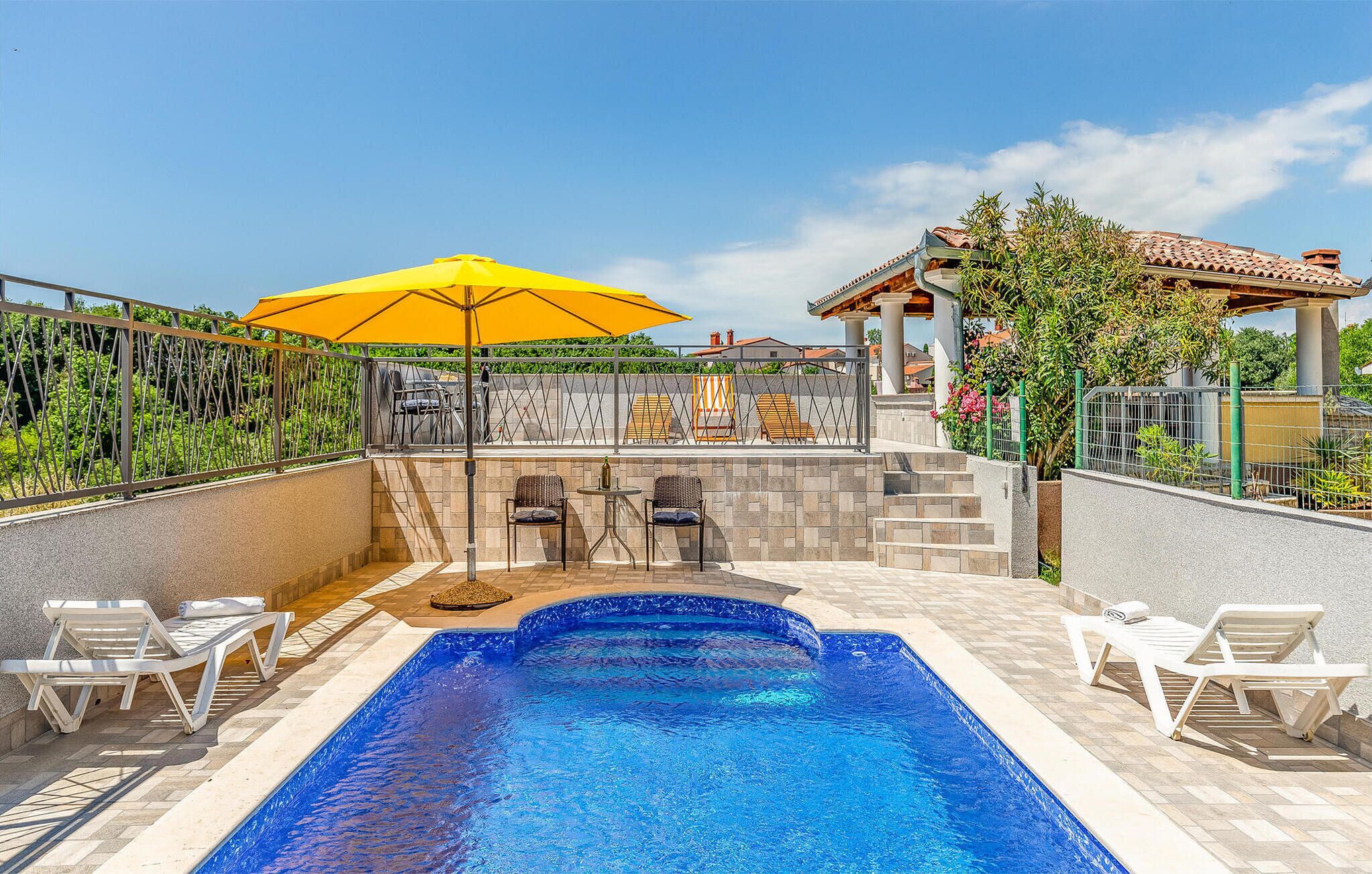 Piscina com espreguiçadeiras e um guarda-sol amarelo, cercada por um muro de pedra e vegetação.