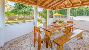 Een overdekt terras met een houten tafel en bankjes, omgeven door stenen vloeren en witte muren.