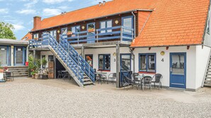 Un bâtiment à deux étages, doté d’un toit recouvert de tuiles rouges, d’un escalier bleu et d’un espace extérieur aménagé pour s’asseoir.