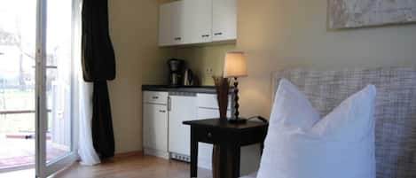 Une chambre à coucher avec un lit, une table de chevet, une lampe, une fenêtre avec des rideaux et, au fond, une petite cuisine.