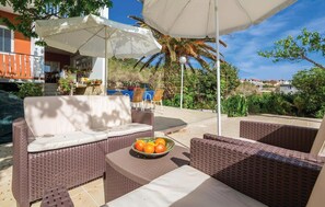 Relax in a serene outdoor seating area with wicker sofas and armchairs, surrounded by a lush garden and shaded by umbrellas.