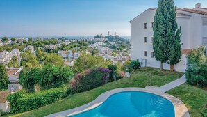 A swimming pool with a curved edge, surrounded by a well-maintained lawn and various plants. In the background, there is a view of a residential area with white buildings and a hill with trees. - Beach front apartment in Mijas Costa (Mijas Costa)