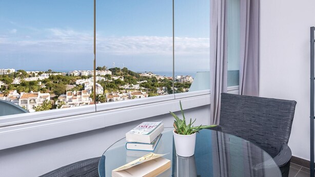 A balcony with a glass table, a potted plant, and books. A cityscape with white buildings and greenery is visible through the window. - Beach front apartment in Mijas Costa (Mijas Costa)