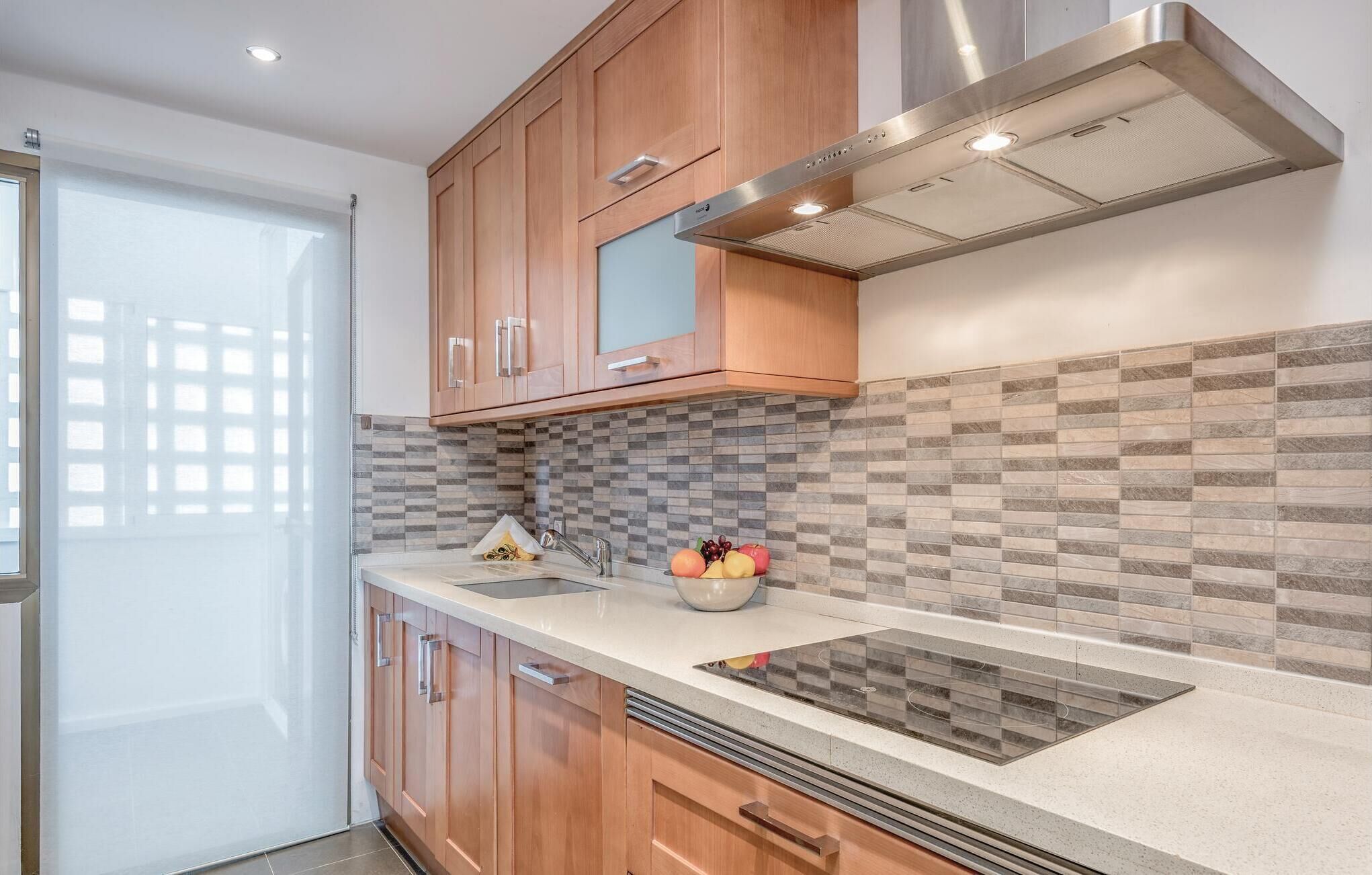 A modern kitchen with wooden cabinets, a stainless steel range hood, a tiled backsplash, and a countertop with a sink and fruit bowl.