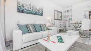 A modern living room with a white sofa, a coffee table with decorative items, a painting on the wall, and a dining area with white chairs and a glass table. - Beach front apartment in Mijas Costa (Mijas Costa)